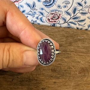 May include: A silver ring featuring an oval-shaped purple gemstone set within a decorative silver bezel. The ring is displayed on a wooden surface.