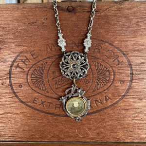 Könnte beinhalten: Eine silberne Kettenkette mit einem großen, verzierten Anhänger mit floralem Design und einem kleineren Anhänger mit einem gerahmten Foto eines jungen Mädchens. Die Kette wird auf einer Holzkiste mit dem aufgedruckten Text "The Monarch Extra" präsentiert.