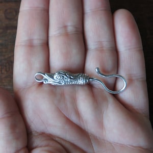 Puede incluir: Un cierre de metal plateado con un diseño detallado de cabeza de pájaro y un gancho en el otro extremo.