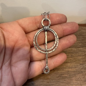 Puede incluir: Un collar con colgante plateado con textura martillada. El colgante presenta dos círculos concéntricos, una barra vertical y un pequeño círculo en la parte inferior. El collar se sostiene en una mano sobre una superficie de madera.
