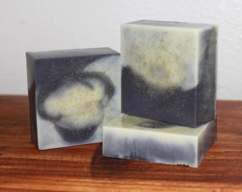 Puede incluir: Tres jabones cuadrados hechos a mano. Los jabones tienen un diseño en capas con una base azul oscuro, un centro blanco y motas doradas. Los jabones están apilados sobre una superficie de madera.