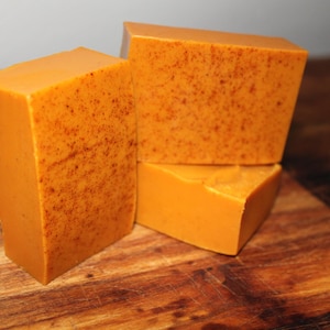 May include: Three bars of orange-coloured soap with a speckled texture. The soap is stacked on a wooden surface.