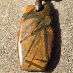 May include: A brown and gold necklace with a large, rectangular pendant made of a patterned stone. The pendant is attached to the necklace with a copper-colored clasp.