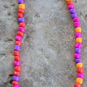 May include: A colorful beaded necklace with square beads in shades of red, orange, purple, and pink. The necklace is displayed on a textured gray surface. The beads are approximately 0.25 inches in size.