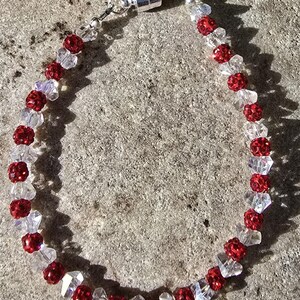 May include: A beaded bracelet featuring alternating red and clear crystal beads. The red beads have a textured, sparkly appearance. The bracelet is secured with a silver magnetic clasp. The jewelry is displayed on a textured, gray surface.
