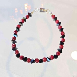 May include: A beaded bracelet featuring alternating red, spherical beads and black, triangular beads. The bracelet has a silver-colored magnetic clasp.