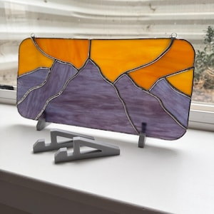 May include: Stained glass panel depicting a mountain range under a sunset. The mountains are purple, and the sky is orange and yellow. The panel is rectangular with rounded corners and is displayed on a white surface with two gray stands.