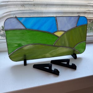 May include: Stained glass panel depicting a landscape with rolling green hills, a yellow sun, and a blue sky. The panel is rectangular with rounded corners and comes with two black stands.