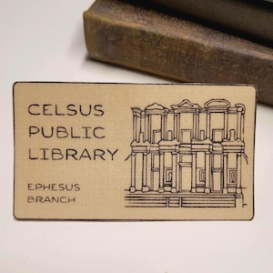 May include: A tan-colored card with black text and a black line drawing of the Celsus Library in Ephesus. The text reads "Celsus Public Library Ephesus Branch".