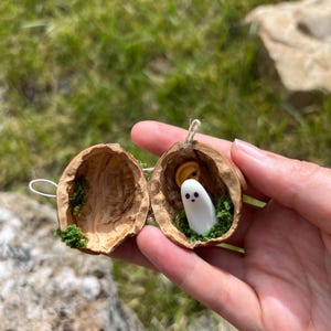May include: A split walnut shell ornament featuring a small white ghost figurine with black eyes and a mouth, nestled inside with green moss and a yellow circular accent. The two halves are connected by a thin string for hanging as a decoration.
