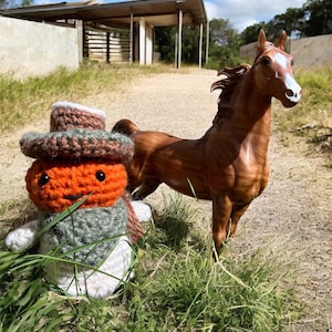 May include: A brown horse figurine stands near a handmade crochet doll with an orange head and a hat. The doll has a green and white body. The scene is outdoors with a building in the background.