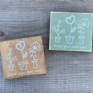 May include: Two square wooden signs featuring white line art of three potted flowers and the text "Grow your own way." One sign has a natural brown wood finish, while the other is painted a muted sage green with visible wood grain texture.