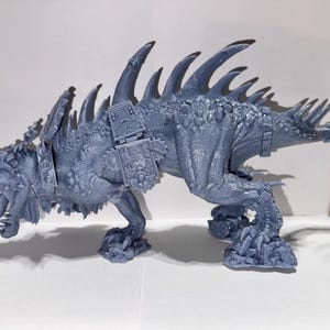 May include: A detailed, grey fantasy creature figurine with a spiky back, horns, and a long tail. The figurine has armour-like details and is posed on all fours. A wooden ruler is in the background.