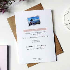 May include: A white RSVP card with a pink and white border. The card has a photo of a couple in a scenic location. The text on the card reads "RSVP", "Please let us know if you can join us by 1st January 2024", "Joe & Alex, 34 Trinity Close, Essex, CM7 2TH", "Alternatively text 07851164485", "We hope you can join us on our special day", "DITSY CHIC", "MADE IN BRISTOL".