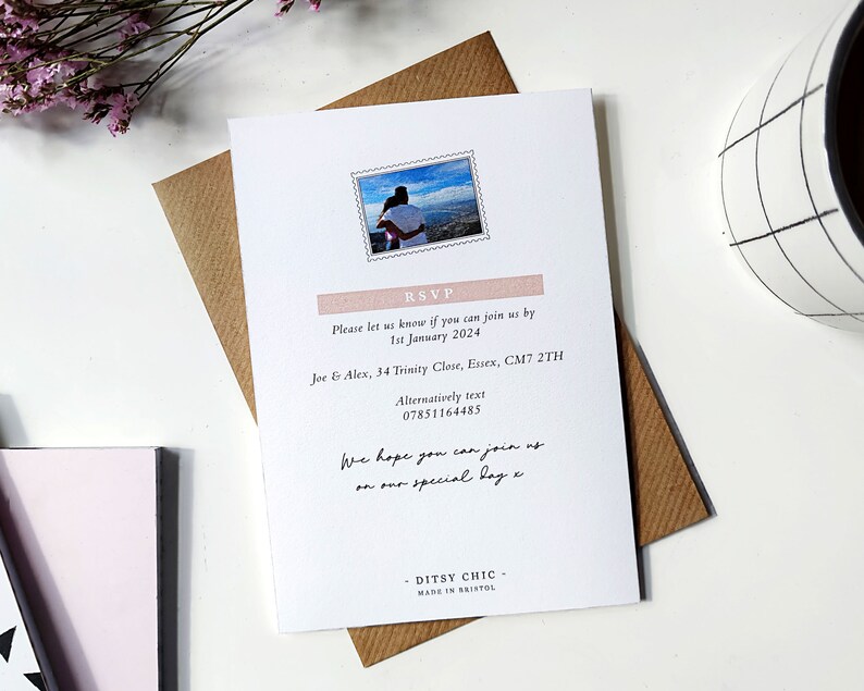 May include: A white RSVP card with a pink and white border. The card has a photo of a couple in a scenic location. The text on the card reads "RSVP", "Please let us know if you can join us by 1st January 2024", "Joe & Alex, 34 Trinity Close, Essex, CM7 2TH", "Alternatively text 07851164485", "We hope you can join us on our special day", "DITSY CHIC", "MADE IN BRISTOL".