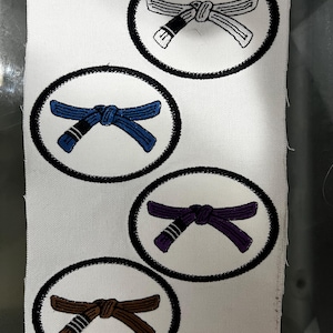 Könnte beinhalten: Vier gestickte Patches mit verschiedenen farbigen Gürteln in einem Kreis. Die Gürtel sind weiß, blau, lila und braun.