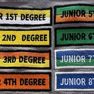 Può includere: Una collezione di toppe ricamate rettangolari con bordo bianco, che riportano la scritta "JUNIOR" seguita da un numero e "DEGREE". Le toppe presentano diverse combinazioni di colori, tra cui giallo, arancione, verde, blu, viola e marrone.
