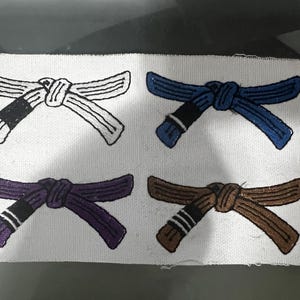 Puede incluir: Cuatro parches bordados que representan cinturones de artes marciales de diferentes colores. Los cinturones son blanco, azul, morado y marrón. Cada cinturón está atado en un nudo y tiene una franja negra en el centro.