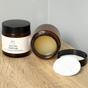 Puede incluir: Dos frascos de vidrio marrón con tapas negras, uno en posición vertical y el otro acostado de lado. El frasco vertical tiene una etiqueta blanca con texto. Una tapa blanca y una tapa negra están al lado del frasco de lado. Los frascos están sobre una superficie de madera.