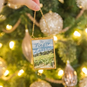 May include: A small, rectangular ornament with a gold-colored frame and chain is held up against a Christmas tree. The ornament features a landscape painting of The Finger Lakes, New York. The background is a blurred Christmas tree with gold and silver ornaments.
