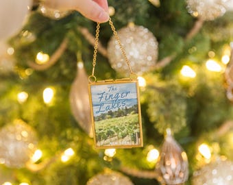 May include: A small, rectangular ornament with a gold-colored frame and chain is held up against a Christmas tree. The ornament features a landscape painting of The Finger Lakes, New York. The background is a blurred Christmas tree with gold and silver ornaments.