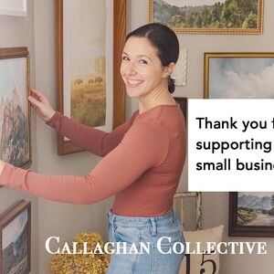May include: A woman smiles while hanging a framed landscape painting on a wall. The painting has a gold frame. The text "Thank you for supporting my small business!" is visible. The business name "Callaghan Collective" is also visible.