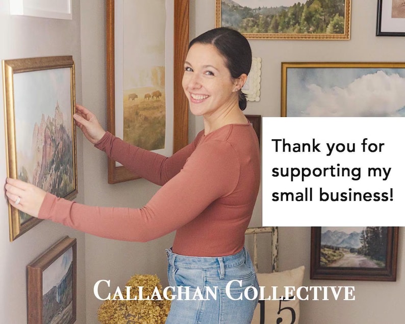 May include: A woman smiles while hanging a framed landscape painting on a wall. The painting has a gold frame. The text "Thank you for supporting my small business!" is visible. The business name "Callaghan Collective" is also visible.