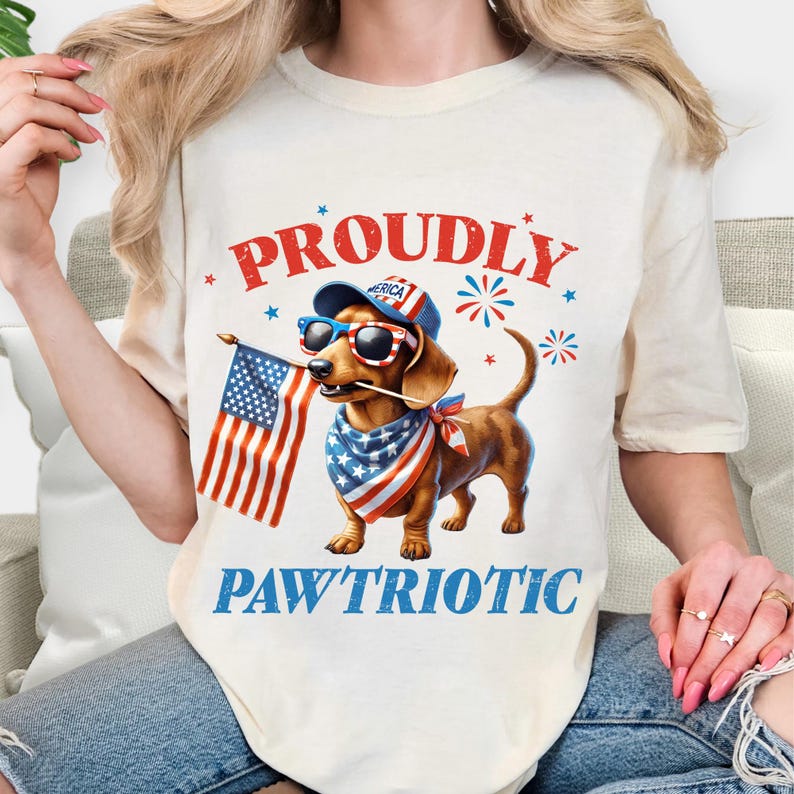 May include: Cream-colored t-shirt featuring a dachshund wearing sunglasses, a patriotic bandana, and a hat with the word "America." The dog holds an American flag. The words "PROUDLY" and "PAW'TRIOTIC" are printed above and below the dog.