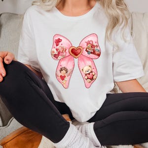 May include: A white t-shirt with a pink bow design. The bow has a heart in the center and is decorated with Valentine's Day themed images, including a teddy bear, an angel, and a red truck.