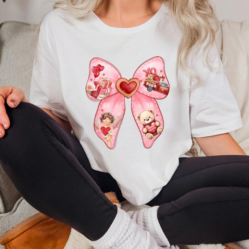 May include: A white t-shirt with a pink bow design. The bow has a heart in the center and is decorated with Valentine's Day themed images, including a teddy bear, an angel, and a red truck.