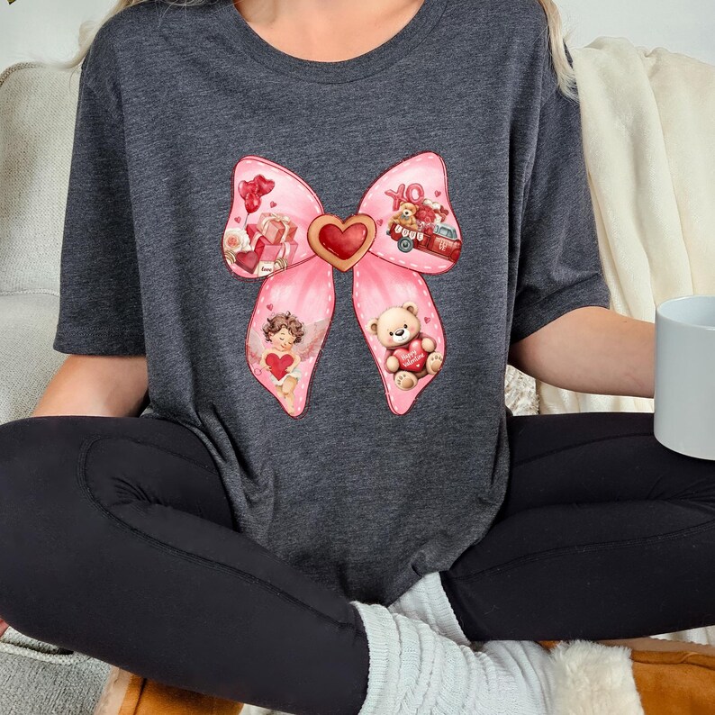 May include: A gray t-shirt with a pink bow design. The bow has a heart in the center and is decorated with images of a teddy bear, an angel, and a red truck with gifts.