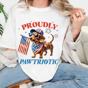 May include: White t-shirt featuring a dachshund wearing sunglasses, a patriotic bandana, and a hat with the word "America." The dog holds an American flag, with the words "Proudly Paw'triotic" in red and blue.