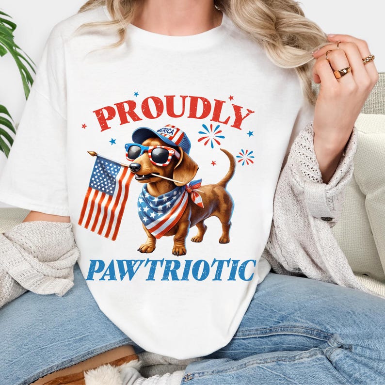 May include: White t-shirt featuring a dachshund wearing sunglasses, a patriotic bandana, and a hat with the word "America." The dog holds an American flag, with the words "Proudly Paw'triotic" in red and blue.