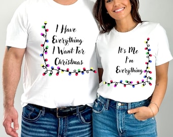 May include: White t-shirts with festive text. One shirt says "I Have Everything I Want For Christmas" and the other says "It's Me I'm Everything". Both shirts feature a border of colourful Christmas lights.