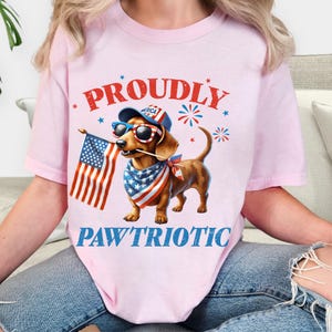 May include: Pink t-shirt with a patriotic dachshund illustration. The dog wears sunglasses, a bandana, and a hat, holding an American flag. Text reads "PROUDLY" and "PAWTRIOTIC" in red and blue.