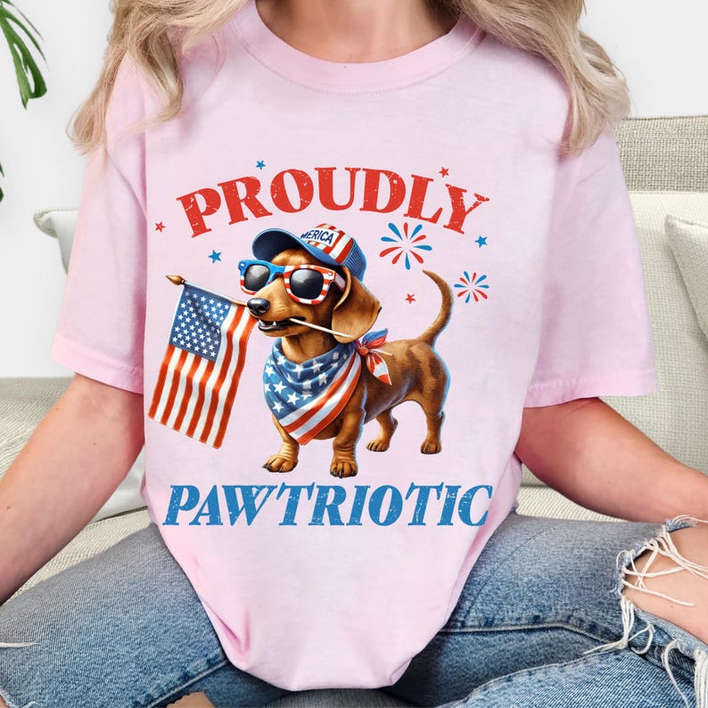 May include: Pink t-shirt with a patriotic dachshund illustration. The dog wears sunglasses, a bandana, and a hat, holding an American flag. Text reads "PROUDLY" and "PAWTRIOTIC" in red and blue.