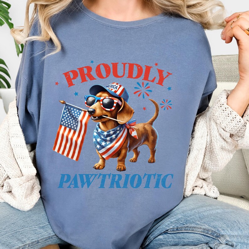 May include: A light blue t-shirt with the words "PROUDLY PAWTRIOTIC" in red and blue. The shirt features a cartoon dachshund wearing sunglasses, a bandana, and a hat, holding an American flag. Patriotic theme.