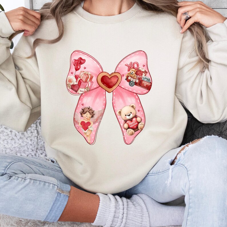 May include: A cream-colored sweatshirt with a pink bow design. The bow is decorated with hearts, a teddy bear, an angel, and a red truck with the words "XO" on the side.