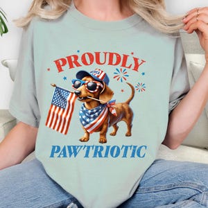 May include: Light blue t-shirt featuring a dachshund wearing sunglasses, a bandana, and a hat, holding an American flag. The shirt has the words "PROUDLY" and "PAWTRIOTIC" in red and blue.