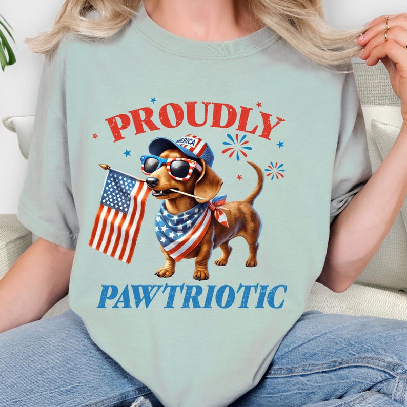 May include: Light blue t-shirt featuring a dachshund wearing sunglasses, a bandana, and a hat, holding an American flag. The shirt has the words "PROUDLY" and "PAWTRIOTIC" in red and blue.