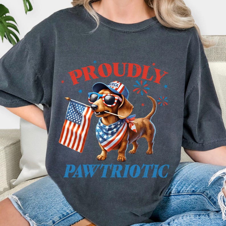 May include: Dark gray t-shirt featuring a dachshund wearing sunglasses, a patriotic bandana, and a hat with the word "America." The dog is holding an American flag. The words "Proudly" and "Pawtriotic" are printed on the shirt in red and blue.