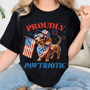 May include: Black t-shirt with a patriotic dachshund graphic. The dog wears sunglasses, a hat, and a bandana, holding an American flag. The words "PROUDLY" and "PAW'TRIOTIC" are printed above and below the dog.