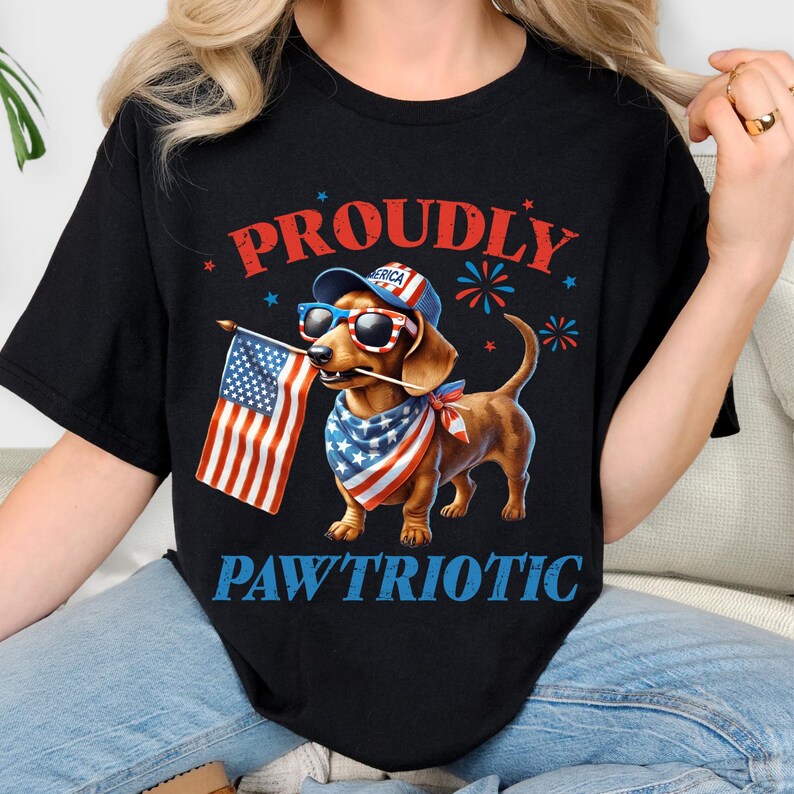May include: Black t-shirt with a patriotic dachshund graphic. The dog wears sunglasses, a hat, and a bandana, holding an American flag. The words "PROUDLY" and "PAW'TRIOTIC" are printed above and below the dog.