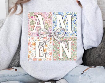 Puede incluir: Una sudadera gris con un diseño floral que deletrea la palabra "AMEN" en letras blancas. Un lazo blanco está en el centro del diseño.