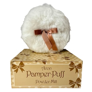以下が含まれることがあります： ヴィンテージのAvon Pamper-Puffパウダーミット。ふわふわの白いパウダーパフには、茶色のサテンのリボンが付いています。「Avon Pamper-Puff Powder Mitt」の文字が前面に印刷された箱の上にパウダーパフが置かれています。箱は、リボンのアクセントが付いた薄茶色の背景です。