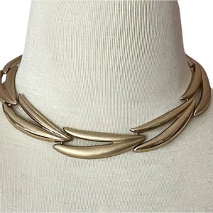 May include: A gold-toned metal choker necklace with a geometric design. The necklace is made of multiple curved pieces that form a continuous loop.