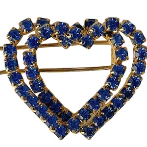 May include: A gold-toned hair clip in the shape of a double heart. The hearts are outlined with small, square, blue gemstones. The clip has two gold-colored metal prongs for securing it in the hair.