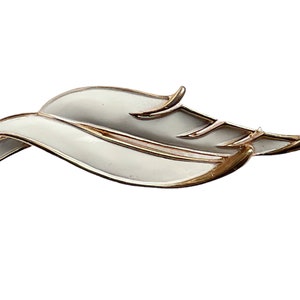 May include: A gold-toned leaf-shaped brooch with white enamel. The brooch has a delicate, flowing design.