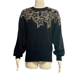 May include: Black long-sleeve sweater with a crew neck. The sweater features a design of colorful stars with sequins and beads across the shoulders and upper chest. Small white dots are scattered across the upper portion of the sweater.
