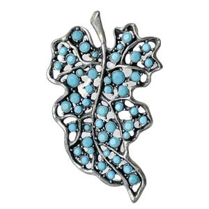May include: A silver-toned leaf-shaped brooch with a filigree design. The brooch is adorned with numerous round, light blue gemstones, creating a striking contrast against the metal. The leaf has a detailed, openwork pattern.